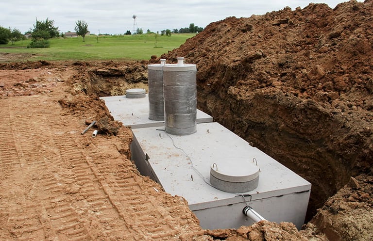 Concrete septic tank installation in an excavated pit with soil mounds surrounding the construction site in a rural area.
